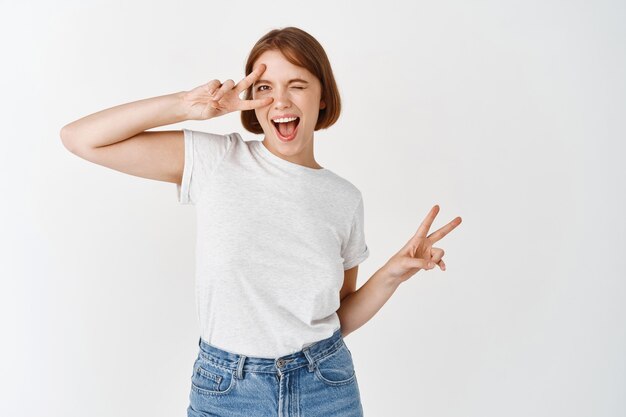 Belle fille caucasienne avec un sourire candide, montrant un signe v sur un œil clignotant et l'air joyeux, debout en jeans et t-shirt contre un mur blanc