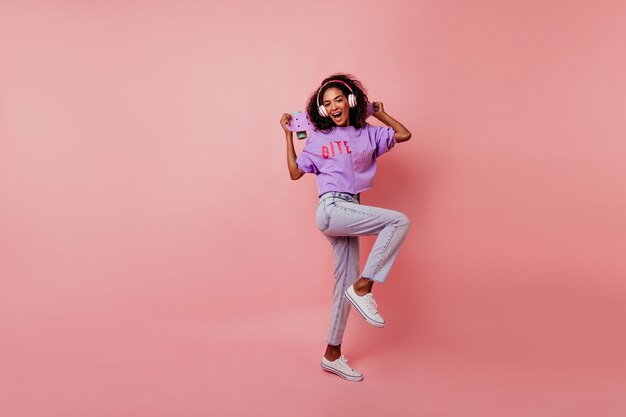 Belle fille en chaussures blanches dansant en studio tout en écoutant de la musique. Portrait en pied d'une femme africaine raffinée avec planche à roulettes refroidissant sur rose.