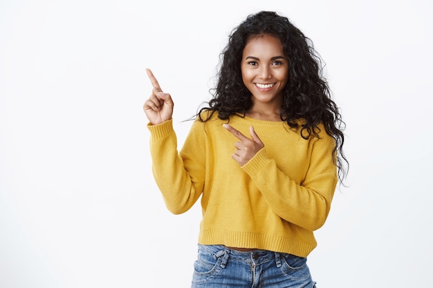 Photo gratuite belle fille confiante avec une coiffure frisée, regarde la caméra avec assurance et sourire impertinent, pointant le coin supérieur gauche, présente un produit étonnant, donne des recommandations