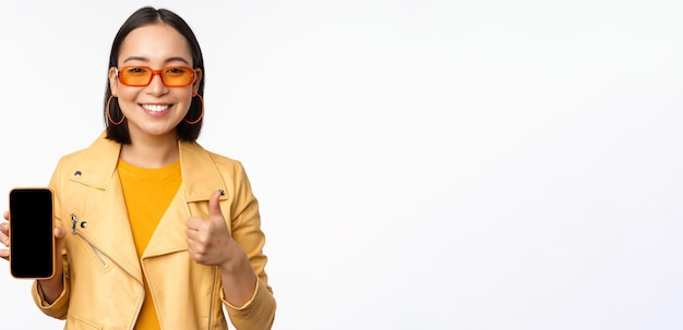 Belle fille coréenne femme asiatique à lunettes de soleil montrant l'interface de l'application smartphone pouce levé recommandant l'application de téléphone mobile fond blanc