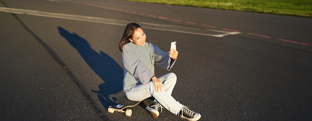 Photo gratuite une belle fille coréenne prend un selfie sur son smartphone prend une photo avec son skateboard en profitant d'une journée ensoleillée