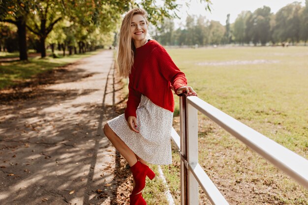 Belle fille dans de belles chaussures rouges et robe blanche s'amusant dans le parc. Belle femme blonde posant de manière ludique près des feuilles mortes jaunes.