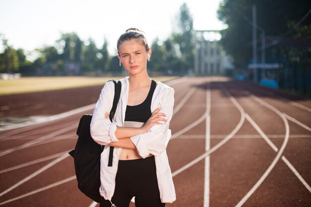 Belle fille dans des écouteurs sans fil avec sac à dos sur l'épaule regardant pensivement à huis clos sur le tapis roulant du stade de la ville