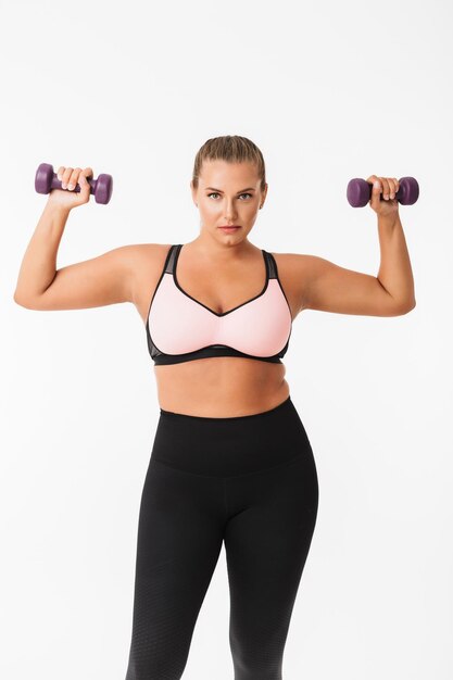 Belle fille dodue en haut sportif et leggings tenant des haltères dans les mains tout en regardant pensivement à huis clos sur fond blanc