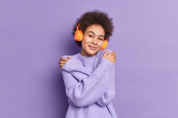 Belle fille du millénaire à la peau sombre embrasse elle-même touche les épaules apprécie la douceur de son nouveau pull écoute une mélodie agréable via des écouteurs stéréo