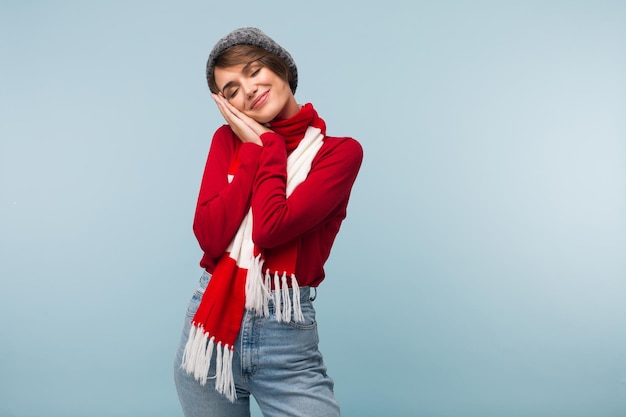 Belle fille en écharpe pull rouge et bonnet tricoté fermant rêveusement les yeux tout en montrant un geste endormi avec les mains sur fond bleu