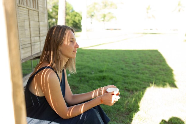 Belle fille est assise près du bungalow et boit du café.