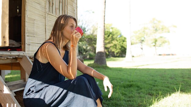 Belle fille est assise près du bungalow et boit du café.