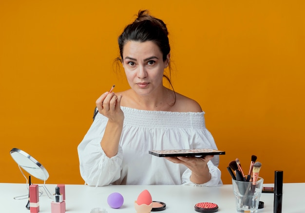 Belle fille est assise à table avec des outils de maquillage tenant un pinceau de maquillage et une palette de fards à paupières à la recherche d'isolement sur le mur orange