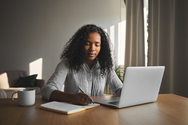 Belle fille étudiante métisse avec une coiffure volumineuse travaillant à la maison dans sa chambre, assise à un bureau en bois, utilisant un ordinateur portable et écrivant dans un cahier. Les gens, la technologie et l'éducation