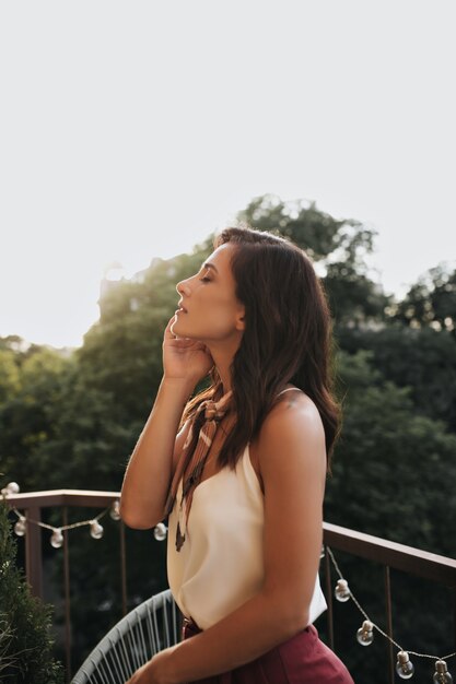 Belle fille en haut blanc et foulard en soie bénéficie d'une journée d'été sur le balcon. Photo de profil de la belle femme en tenue élégante sur la terrasse.