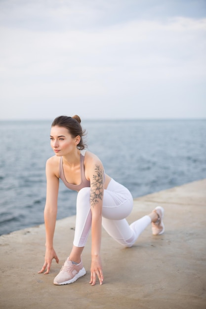 Belle fille en haut sportif et leggings blancs pratiquant le yoga avec vue sur la mer en arrière-plan. Jeune femme regardant pensivement de côté tout en s'entraînant au bord de la mer