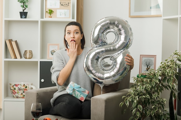 Belle fille le jour de la femme heureuse tenant le ballon numéro huit assis sur un fauteuil dans le salon