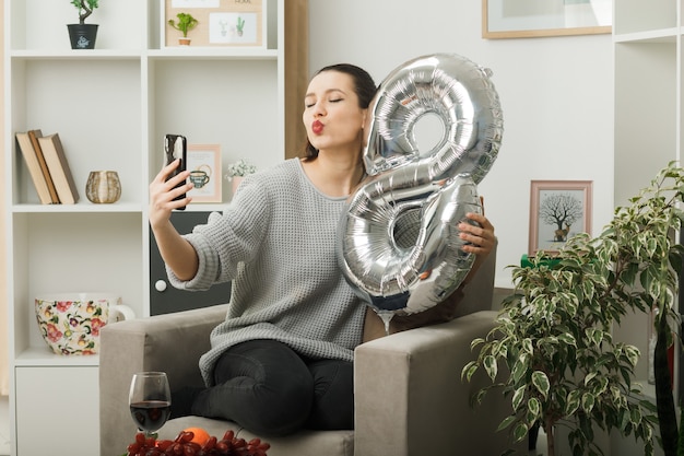 Belle fille le jour de la femme heureuse tenant le ballon numéro huit assis sur un fauteuil dans le salon