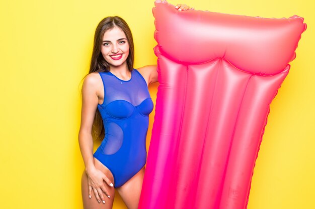 Belle fille joyeuse dans un maillot de bain lumineux avec un matelas pneumatique sur un mur jaune.