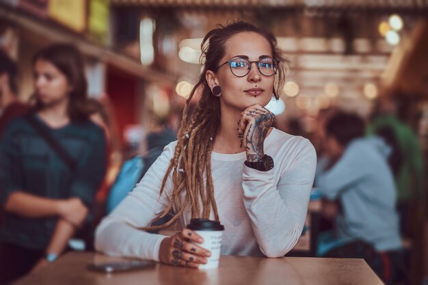 Belle fille joyeuse avec des tatouages et des dreadlocks est assise à l'aire de restauration tout en buvant du café.