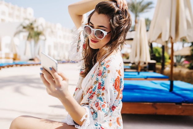 Belle fille en lunettes de soleil blanches à la mode écrivant un message à un ami, tenant ses cheveux noirs. Portrait d'une incroyable jeune femme brune en chemise élégante assis à l'extérieur avec téléphone