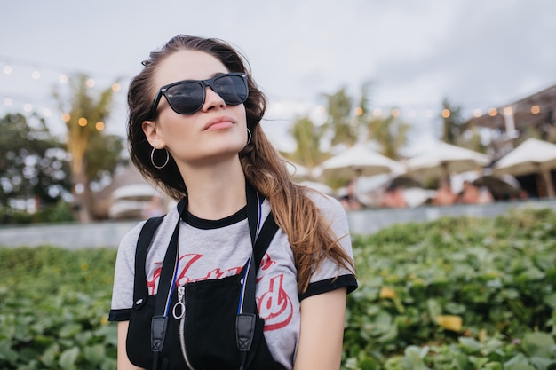 Photo gratuite belle fille en lunettes de soleil noires regardant le ciel sur la nature. tir extérieur d'une femme brune à la mode, passer du temps en plein air en soirée.