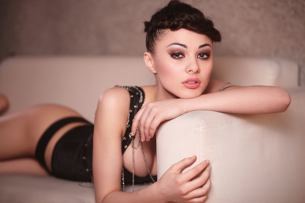 Belle fille de mode en corset noir allongé sur le canapé à l'intérieur maquillage et coiffure lumineux