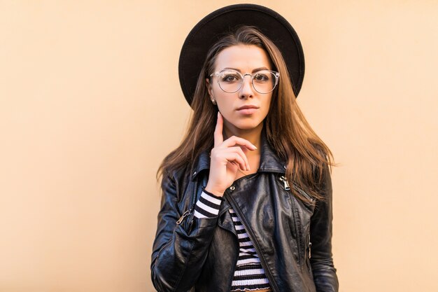 Belle fille de mode sérieuse en veste de cuir et chapeau noir isolé sur un mur jaune clair