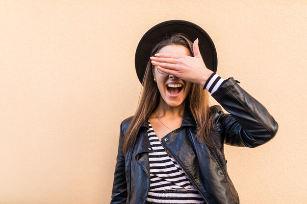 Belle fille de mode en veste de cuir et chapeau noir couvrir son œil de visage avec sa main isolée sur un mur jaune clair