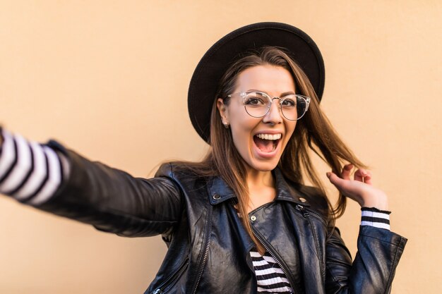 Belle fille de mode en veste de cuir, lunettes transparentes et chapeau noir fait selfie isolé sur mur jaune clair