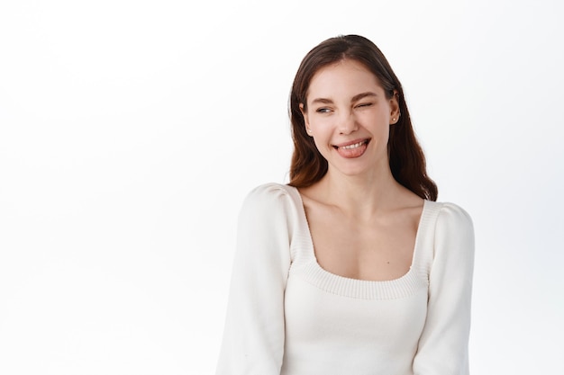 Belle fille modèle clignant de l'œil et montrant la langue, faisant un visage positif idiot, regardant de côté le logo de l'entreprise ou le texte promotionnel, debout contre un mur blanc