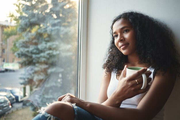 Belle fille mulâtre à la peau sombre dans des vêtements élégants se détendre par la fenêtre à la maison, tenant une grande tasse, profitant d'un café ou d'un thé frais, ayant une expression faciale de rêve, pensant à quelque chose d'agréable