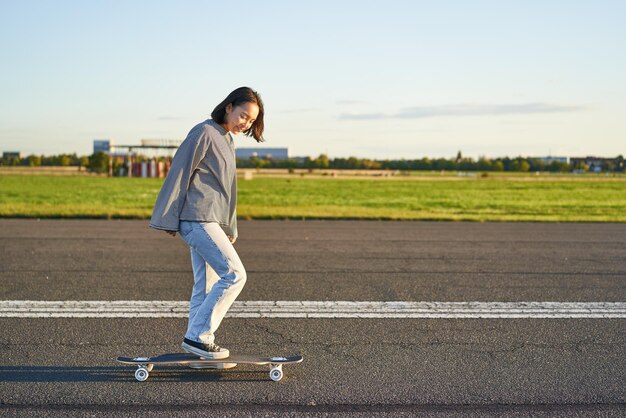 Belle fille de patineur asiatique chevauchant son longboard sur une route déserte ensoleillée jeune femme appréciant son skate