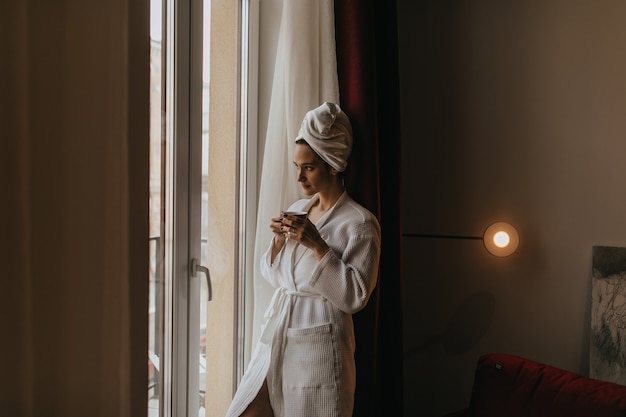 Belle fille en peignoir et serviette sur la tête regarde pensivement par la fenêtre avec une tasse de thé dans ses mains.