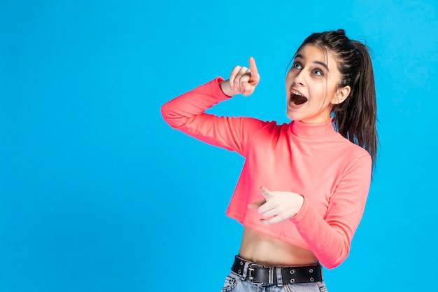 Une belle fille pointant son doigt en l'air et debout sur fond bleu Photo de haute qualité