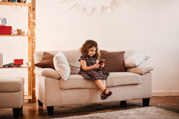 Belle fille préadolescente en robe assise sur un canapé. Plan intérieur d'un petit enfant bouclé posant dans le salon.