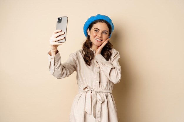 Photo gratuite belle fille prenant selfie sur smartphone en manteau et chapeau mignon, souriant pour la photo et l'air heureux, debout sur fond beige
