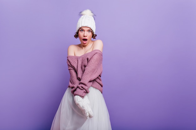 Belle fille en pull violet exprimant l'étonnement isolé sur un mur lumineux. Surpris femme brune aux cheveux courts en jupe blanche debout