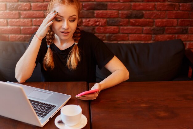 Belle fille de race blanche au gingembre à l'aide d'un ordinateur portable et d'un téléphone intelligent alors qu'il était assis sur un canapé avec une émotion de visage étonnant