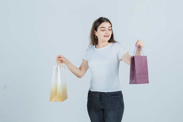 Belle fille regardant des sacs-cadeaux en t-shirt, jeans et à la surprise. vue de face.