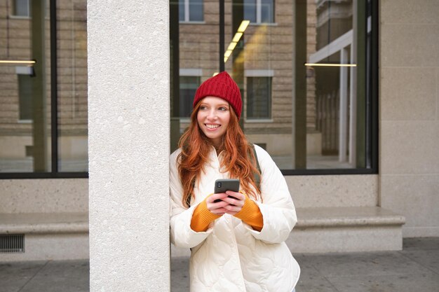 Photo gratuite belle fille rousse avec un smartphone debout dans la rue tenant un téléphone mobile à l'aide d'une application de taxi connec