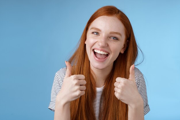 Une belle fille rousse sortante enthousiaste et amusée dit oui, soutient comme une idée géniale, montre l'approbation du pouce levé, recommande le geste d'accord, bon choix, fond bleu debout. Espace de copie