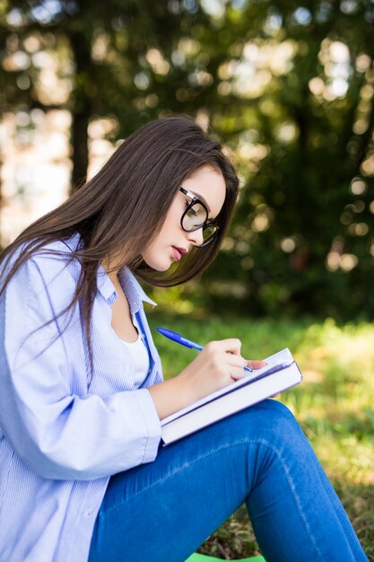 Belle fille sérieuse aux cheveux noirs en veste de jeans et lunettes écrire dans le cahier dans le parc