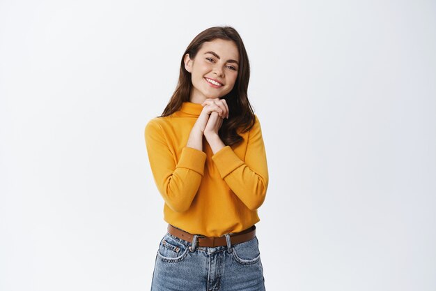 Belle fille souriante à l'air heureuse et reconnaissante, recevant un cadeau et l'admirant, regardant devant elle satisfaite des mains pressées contre la poitrine, mur blanc