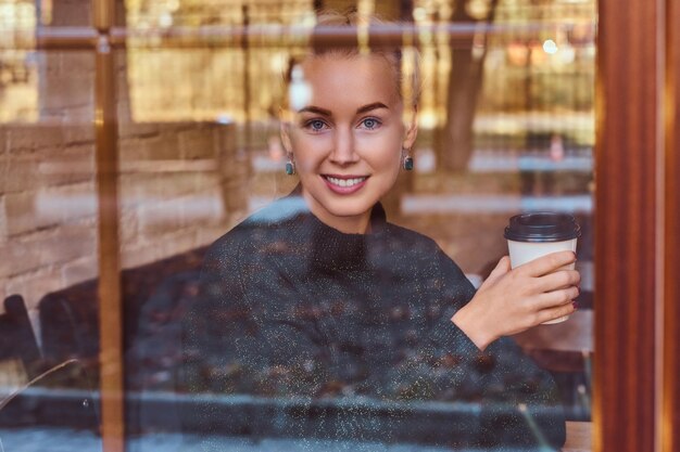 Une belle fille souriante buvant du café dans le café derrière la fenêtre.