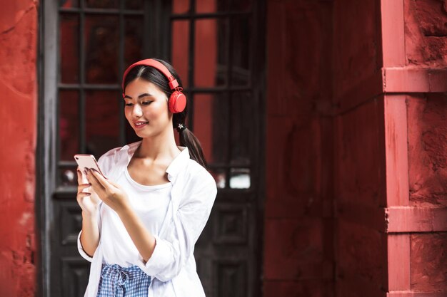 Belle fille souriante en chemise blanche et casque rouge utilisant joyeusement un téléphone portable tout en passant du temps dans la cour