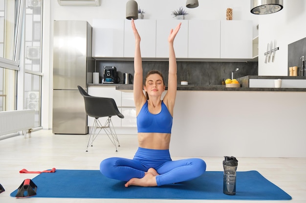 Photo gratuite une belle fille souriante fait du yoga dans son salon en levant les mains et en pratiquant la respiration profonde.