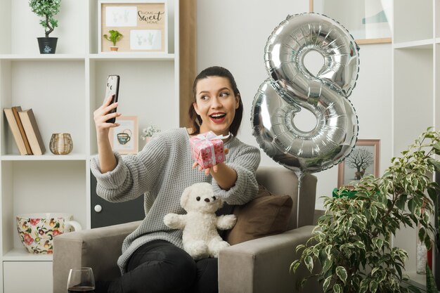 Belle fille souriante le jour de la femme heureuse tenant un cadeau prendre un selfie assis sur un fauteuil dans le salon