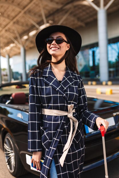 Belle fille souriante à lunettes de soleil et chapeau noir regardant joyeusement de côté près de l'aéroport avec voiture cabriolet sur fond