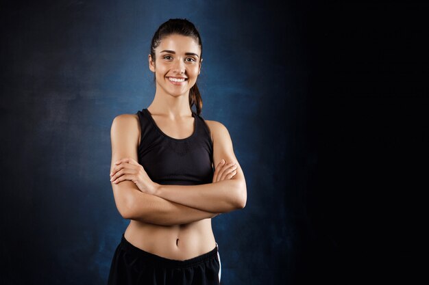 Belle fille sportive posant avec les bras croisés sur un mur sombre.