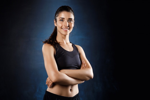 Belle fille sportive posant avec les bras croisés sur un mur sombre.