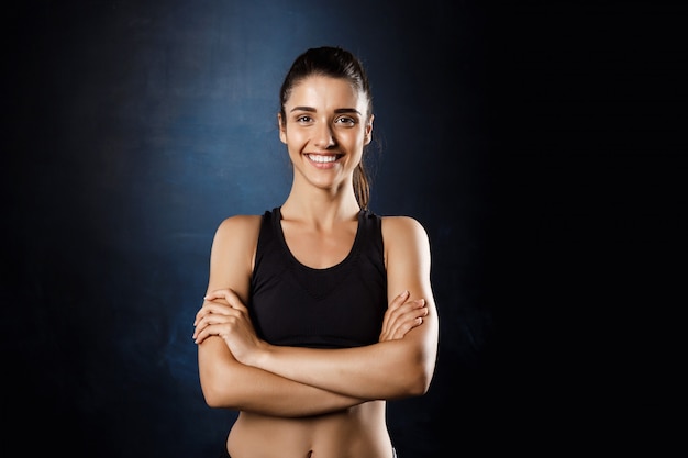 Belle fille sportive posant avec les bras croisés sur un mur sombre.