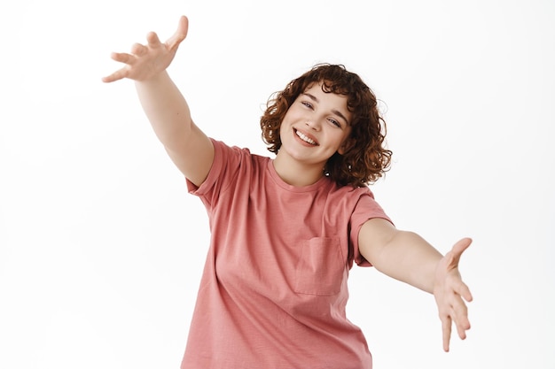 Belle fille sympathique écarte les mains pour se faire un câlin, accueille quelqu'un avec des étreintes chaleureuses et heureuses, tend les mains vers la caméra, debout sur fond blanc