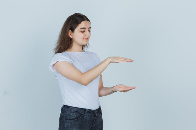 Belle fille en t-shirt, jeans montrant le signe de la taille et l'air heureux, vue de face.